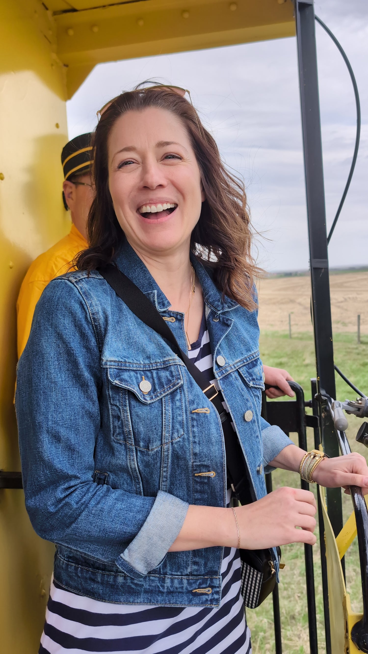 Woman in a denim jacket laughing joyfully outdoors, with prairie fields and a vintage yellow train car in the background — capturing spontaneous, in-the-moment energy.