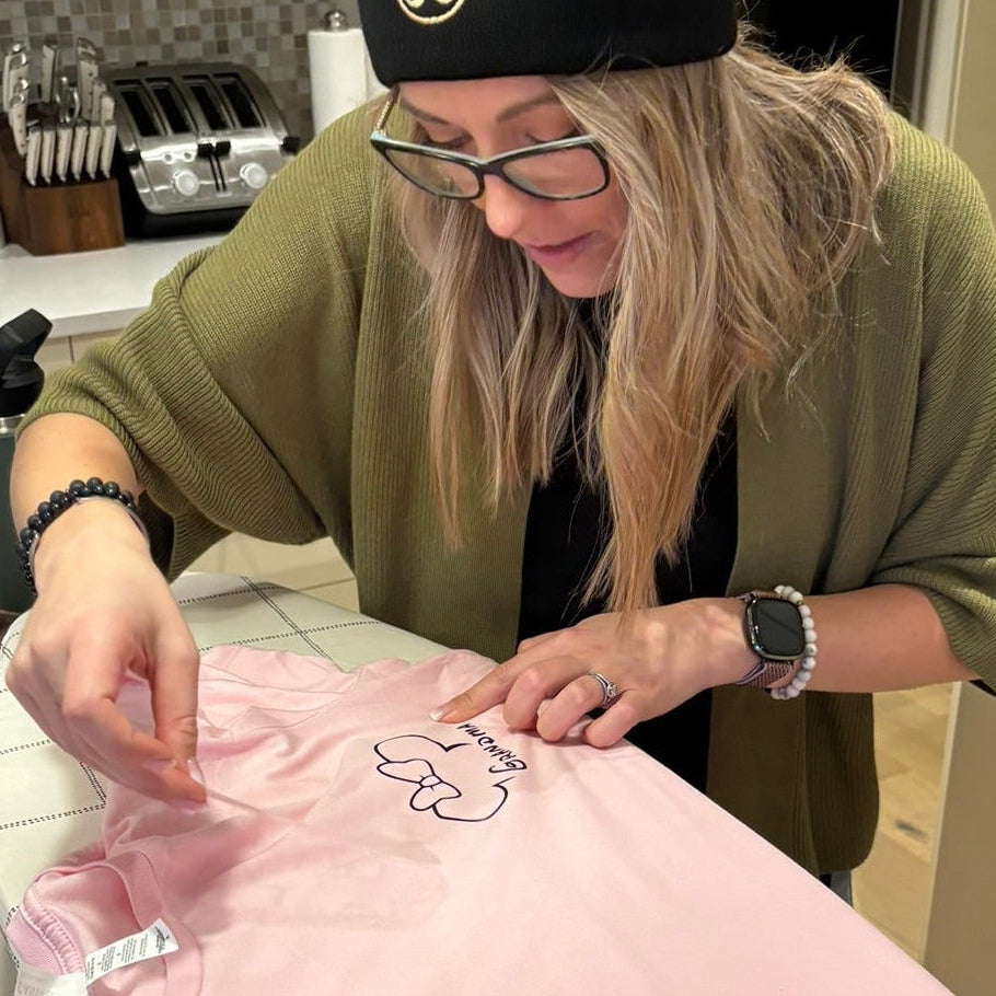 A warm indoor scene showing a woman seated at a worktable carefully hand‑pressing a design onto a soft pink garment, surrounded by crafting tools and materials. Her focused posture, glasses, and relaxed smile create a sense of creativity and personal connection, reflecting the hands‑on, heartfelt work that represents the spirit of the Salt & Spice Collective.