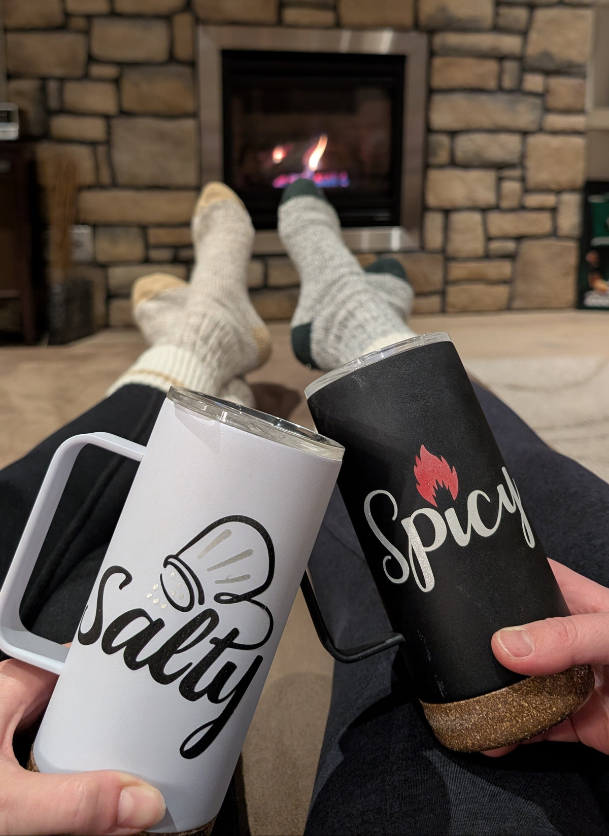 An indoor moment showing two women sitting comfortably by a fireplace, in their comfy, cozy socks, while holding a pair of branded Salt & Spice mugs, creating a warm, relaxed atmosphere that highlights the cozy, inviting spirit behind the Salt & Spice Collective. The soft lighting, casual setting, and personal touch emphasize friendship, connection, and the meaningful beginnings of the brand’s story.