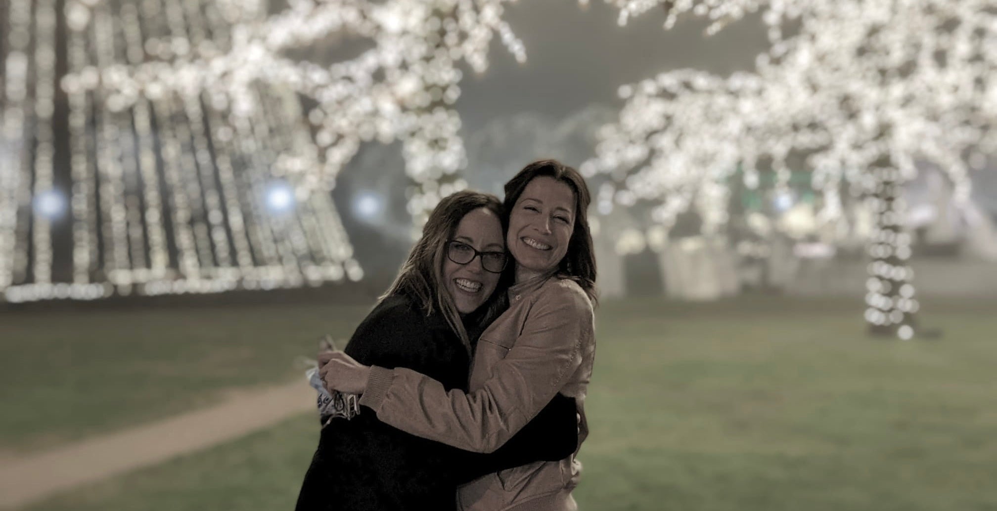 A nighttime outdoor scene showing two women embracing warmly in front of a large display of glowing string lights, standing in a softly lit park area with blurred trees and pathways behind them. Their winter coats and close posture create a feeling of comfort and connection, capturing a heartfelt moment that reflects the deep friendship and supportive bond central to the Salt & Spice Collective.