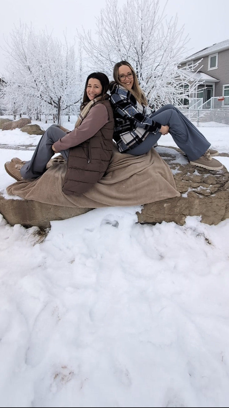 A winter outdoor scene showing two women sitting back‑to‑back on a large snow‑covered rock in a quiet suburban neighborhood, dressed in layered coats, scarves, and boots that create a warm contrast to the snowy surroundings. One woman smiles toward the camera while the other looks out at the landscape, and behind them leafless trees and nearby homes are coated in fresh snow, capturing a peaceful moment of closeness and the deep friendship that inspired the origins of the Salt & Spice Collective