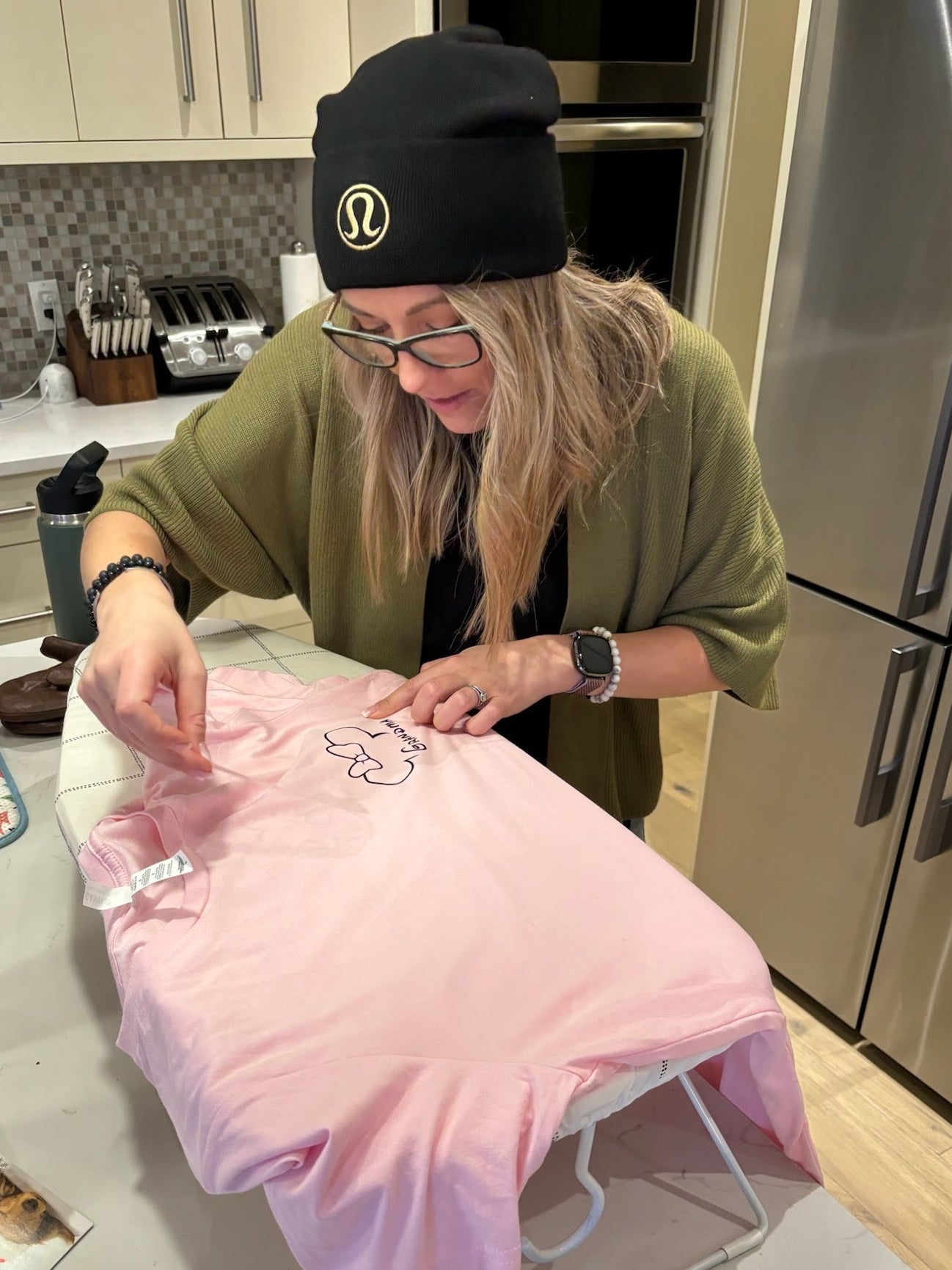 A warm indoor scene showing a woman seated at a worktable carefully hand‑pressing a design onto a soft pink garment, surrounded by crafting tools and materials. Her focused posture, glasses, and relaxed smile create a sense of creativity and personal connection, reflecting the hands‑on, heartfelt work that represents the spirit of the Salt & Spice Collective.